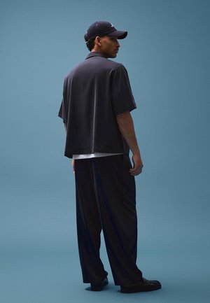 Uomo con cappellino nero, camicia nera a maniche corte larga, pantaloni neri ampi e scarpe nere, in piedi contro uno sfondo blu, di profilo.