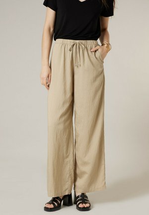 Vrouw draagt beige wijde broek met trekkoord in de taille, zwart kort mouw shirt, zwarte sandalen met hak en gouden armbanden aan haar linker pols.