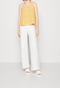 Débardeur floral jaune à fines bretelles, associé à un pantalon large blanc et des sandales compensées blanches. La tenue a une coupe décontractée.