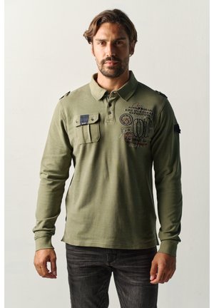 Hombre con camisa tipo polo de manga larga verde oliva con bolsillo en el pecho y texto impreso, de pie contra un fondo liso y claro.