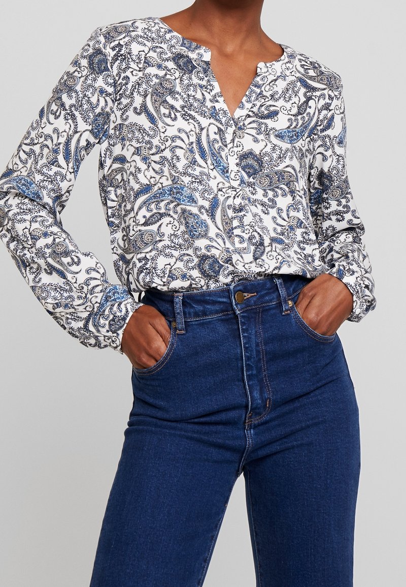 Chemise blanche à manches longues avec un motif paisley bleu, col boutonné et manches cintrées, assortie à un jean taille haute bleu foncé.