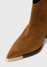 Brune sued bottines med spisse tå og svart såle med gullmetallisk tåhette gravert med logo.