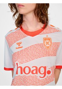 Witte voetbaltrui met oranje accenten. Beschikt over een V-halsontwerp, een textuurpatroon van namen en logo's, waaronder "hoag" en "hummel."