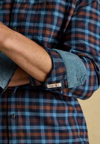 Zils un brūns raibs flaneļa krekls ar džinsa aproci. Iekļauj kabatu un zīmola birku uz piedurknes. Kokvilnas materiāls, ar pogām aizverams dizains.