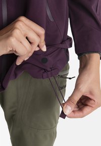 Fioletowa kurtka o teksturowym wykończeniu, z czarnym sznurkiem ściągającym i płaskim czarnym zapięciem, noszona na oliwkowych spodniach.