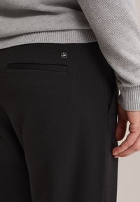 Svarta byxor i texturerat tyg, med en bakficka och en liten metalllogo på midjebandet. Grå stickad tröja synlig ovanför.