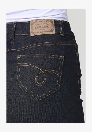 Tumedast sinise denimi teksad kollaste õmblustega, KOROSHI nahkpaat, ning ainulaadne kaarekujuline tasku disain selja peal.