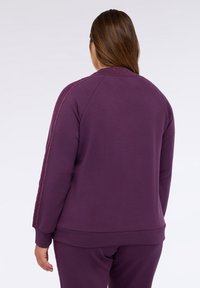 Fialová mikina s dlouhými rukávy, žebrovanými manžetami a límcem, s texturovaným detailem podél ramen. Hladký povrch látky.