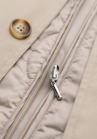 Giacca beige con un grande bottone marrone e una cerniera argentata su un tessuto testurizzato, che mostra cuciture dettagliate e linee morbide.