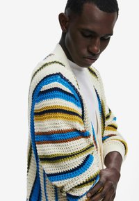 Cardigan lavorato a maglia multicolore con righe blu, gialle e marroni. Presenta una vestibilità comoda e un design aperto con polsini a coste.