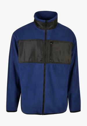 Blå fleecejakke med en sort nylon øverste del, full glidelås og en glidelås-lommer på brystet. Har høy krage og elastiske mansjetter.