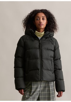 Vrouw met krullend haar in een zwarte pufferjack met capuchon, lichte turtleneck en grijs geruit broek, staande tegen een effen achtergrond.