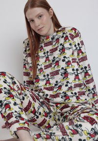Conjunto de pijama de forro polar en color crema que presenta un colorido estampado de Mickey Mouse. Camiseta de manga larga y pantalones con tobillos ajustados. Textura suave y acogedora.