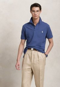 Νεαρός άνδρας με γυαλιά, μπλε polo μπλούζα, μπεζ πλισέ παντελόνι, καφέ ζώνη και ρολόι χειρός, που στέκεται με το ένα χέρι στην τσέπη.