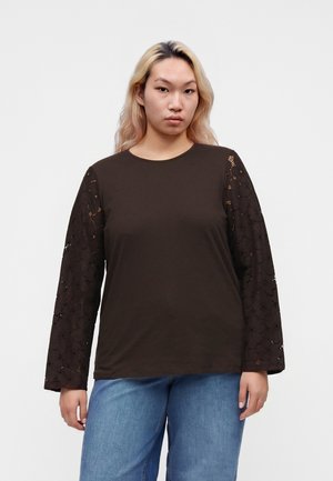 Jeune femme aux longs cheveux blonds porte un haut noir à manches en dentelle et un jean bleu sur un fond clair uni.