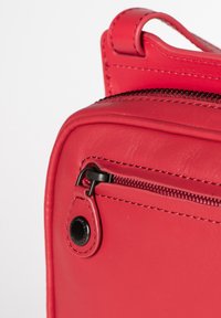 Detail einer roten Ledertasche mit einem Reißverschluss, schwarzem Beschlag und polierter Textur, mit abgerundetem oberen Teil und genähten Akzenten.