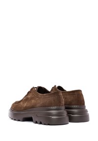 Chaussures en daim marron à lacets avec des semelles en caoutchouc noir épais et des motifs texturés, présentées sous un angle arrière à un quart sur fond blanc.