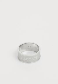 Bague en métal argenté avec un bandeau texturé et le logo gravé "LACOSTE". Diamètre intérieur marqué "62". Finition lisse sur la surface intérieure.
