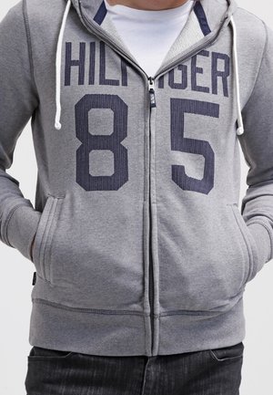 Personne portant un sweat à capuche gris zippé avec "HILFIGER 85" imprimé en grandes lettres et chiffres bleu marine sur le devant, mains dans les poches.