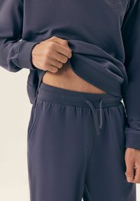 Ciemnofioletowa bluza i dopasowane dresy wyróżniają się miękkim materiałem, elastycznym pasem w talii oraz regulowanym sznurkiem, a także posiadają boczne kieszenie.