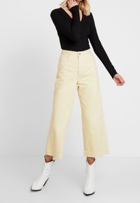 Vaaleankeltaiset leveälahkeiset housut korkea vyötärö, joissa on etutaskut, nappikiinnitys ja sileä puuvillapinta. Yhdistetty mustaan toppiin.