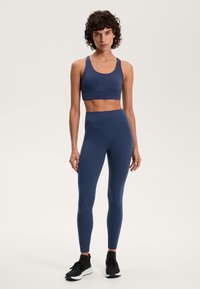 Sötétkék sportmelltartó és leggings szett rugalmas anyagból. A melltartón széles pánt található, a leggings magas derékkal rendelkezik. Fekete sportcipők egészítik ki a megjelenést.