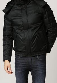 Chaqueta acolchada negra con cuello alto y capucha. Incluye cierre de cremallera y broches, puños elásticos y bolsillos laterales. Tejido liso.