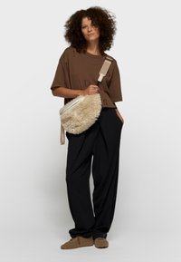 Mujer con cabello rizado que lleva una camiseta marrón extragrande, pantalones anchos negros, zapatillas marrones de peluche y sostiene un bolso cruzado beige y esponjoso.