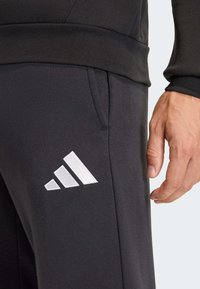 Μαύρο αθλητικό παντελόνι από μαλακό υλικό. Διαθέτει μεγάλο λευκό λογότυπο Adidas και πλαϊνές τσέπες. Το σχέδιο περιλαμβάνει εφαρμοστό ζωνάκι στη μέση και στενές προς τα κάτω γραμμές στα πόδια.