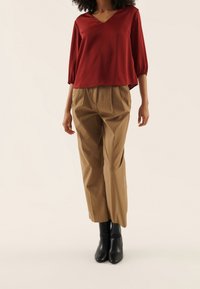 Rotes Satinoberteil mit V-Ausschnitt und drei Viertel Puffärmeln, kombiniert mit tanfarbigen, hoch taillierten, plissierten Hosen und schwarzen Ankle Boots.