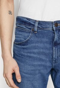 Jeans de mezclilla azul con cinco bolsillos, costuras visibles y cierre tradicional de botón y cremallera. Textura suave y diseño de tiro medio.