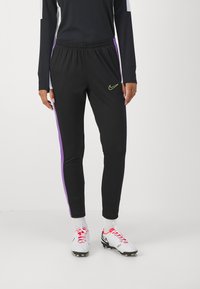 Pantalones deportivos negros con franjas laterales moradas, cintura elástica y un pequeño logotipo de Nike amarillo en el muslo. Material suave y elástico.