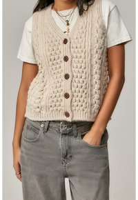Krémová pletená vesta s výstřihem do V, zdobená strukturovanými vzory a pěti hnědými knoflíky, nošená přes bílé tričko a v kombinaci se šedými džíny.