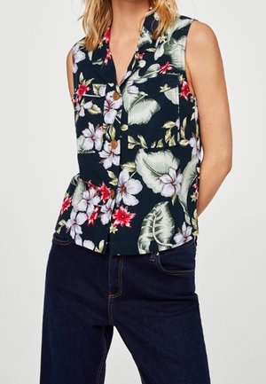 Mouwloos marineblauw blouse met knopen en bloemenprint in rood, wit en groen, gedragen met donker denim broek.