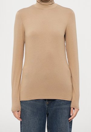Haut beige ajusté à manches longues avec col roulé porté avec un jean denim bleu sur une personne devant un fond clair uni.