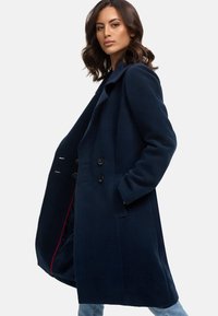 Marineblauwe wollen jas met een geknipte kraag, dubbelzijdige knopen, zijzakken en een gestructureerde afwerking. Het interieur heeft een rode accentkleur.