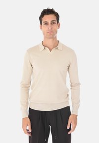 Maglione polo beige a maniche lunghe con design a scollo a V, polsini a costine e struttura aderente, esposto contro uno sfondo bianco uniforme.