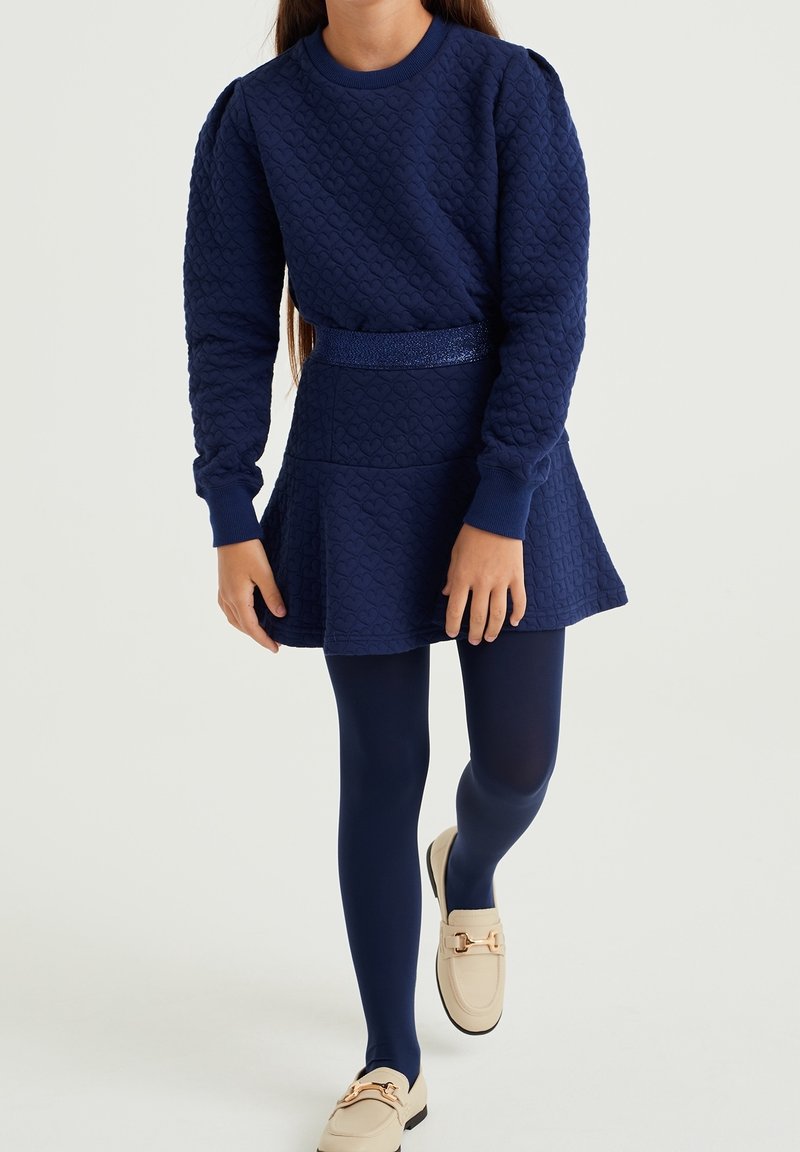Fille portant une robe texturée bleu marine à manches longues, des leggings assortis et des mocassins crème faciles à enfiler avec des détails dorés.