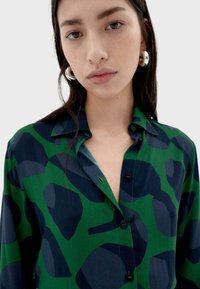Camisa con patrón verde y azul marino, de cuello abotonado, con formas irregulares y una textura de tela suave y ligera.