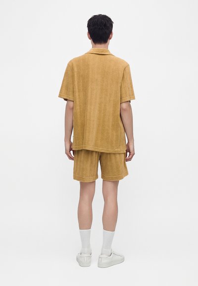 Homme portant une chemise à manches courtes en éponge beige ample et un short assorti, chaussettes blanches et baskets blanches, debout de dos.