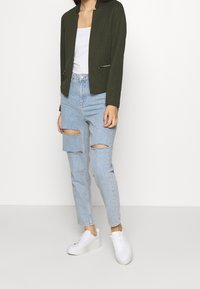 Olivgrön kavaj med krage, i kombination med ljusblå slitna jeans och vita sneakers. Minimalistisk design med dragkedjedetaljer.