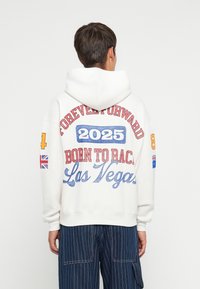 Moletom branco com texto colorido: "PARA SEMPRE PARA A FRENTE," "2025," "NASCIDO PARA CORRER," e "Las Vegas." Inclui bandeiras e acentos em negrito; tecido suave.