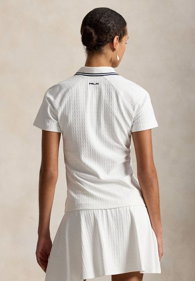 Femeie cu păr închis la culoare prins în coc, purtând o cămașă polo albă texturată cu mâneci scurte și o fustă plisată asortată, văzută din spate pe un fundal neutru.