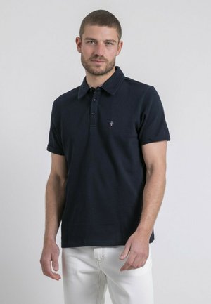 Homme portant un polo bleu marine à manches courtes avec un petit logo sur la poitrine et un pantalon blanc, debout devant un fond clair uni.