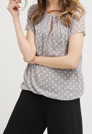 Vrouw draagt een grijs kortarmig blouse met witte stippen en zwarte broek met wijde pijpen, armen gekruist en hand dicht bij het gezicht geheven.