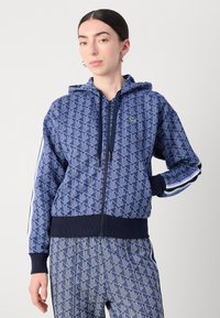 Navyblå zip-up hoodie med geometrisk mönster, dragsko i huva och ribbade muddar. Har logotyp på bröstet och randiga detaljer på ärmarna.