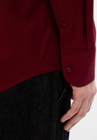Mano appoggiata accanto a jeans scuri, indossando una camicia a maniche lunghe di un rosso scuro con il bottone del polsino visibile.