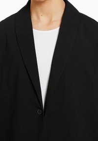 Mustaa bleiseriä, jossa on huppukolkki, yksinäinen nappi ja teksturoitu kangas, kerroksittu valkoisen ribbauspuseron päälle.