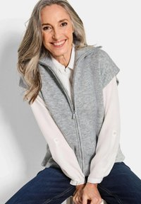 Grauer Strickwesten mit hohem Kragen, Reißverschluss vorne und kurzen Ärmeln, über einem weißen Hemd getragen. Die Textur ist weich und hat ein geripptes Muster.