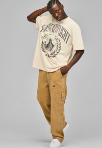 Beige oversized t-skjorte med grafisk design, paret med tan cargo bukser med sidelommer, og hvite sneakers.